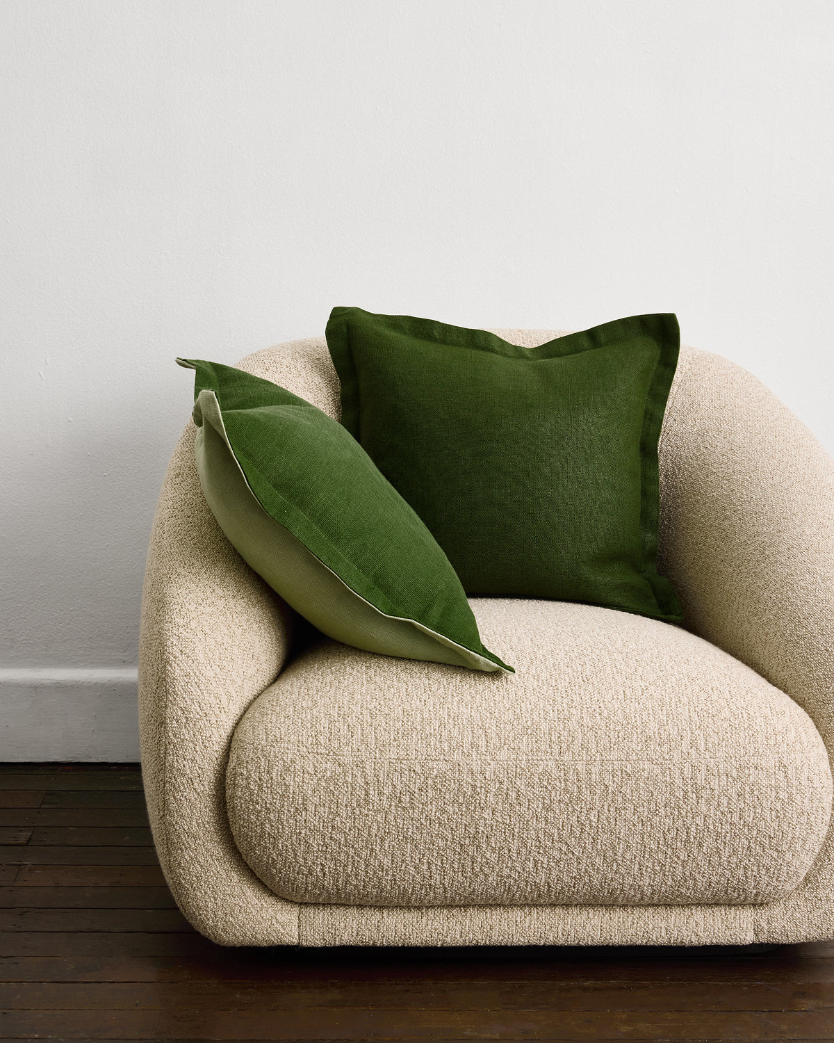 A beige, textured armchair rests on a dark wood floor against a white wall, styled with two Bed Threads Sage & Olive 100% French Flax Linen Cushion Covers—one upright and one diagonal—for a touch of natural elegance.