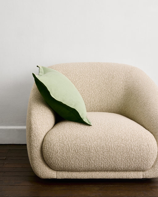 A modern beige upholstered armchair sits on a dark wooden floor against a white wall, with a light green Bed Threads Sage & Olive 100% French Flax Linen Cushion Cover resting on its left arm.