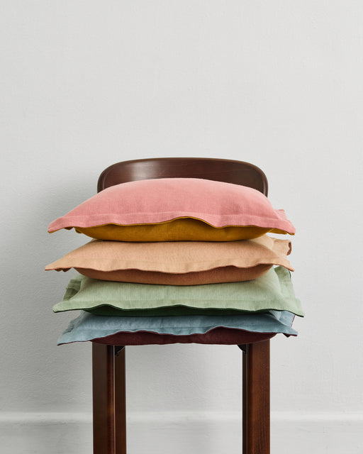 Four Sage & Olive 100% French Flax Linen Cushion Covers by Bed Threads in pink, yellow, green, and light blue are stacked on a dark wooden chair against a plain white wall.