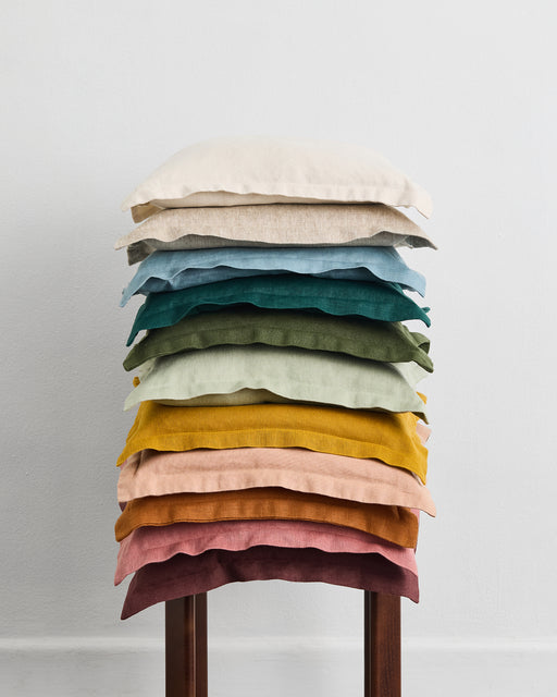 An Oatmeal 100% French Flax Linen Cushion Cover by Bed Threads is arranged neatly on a wooden stool, set against a plain white wall.