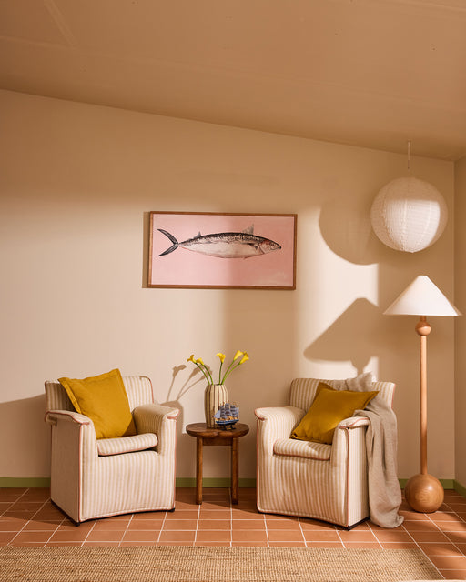A cozy sitting area with two striped armchairs, yellow cushions, a Bed Threads Oatmeal 100% French Flax Linen Throw, a round wooden side table with yellow flowers in a vase, a floor lamp, and a framed fish illustration on pink above.