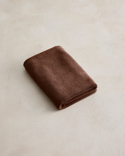 A Bed Threads Espresso Cotton Terry Bath Mat is displayed with a neatly folded brown towel on a light, textured surface.