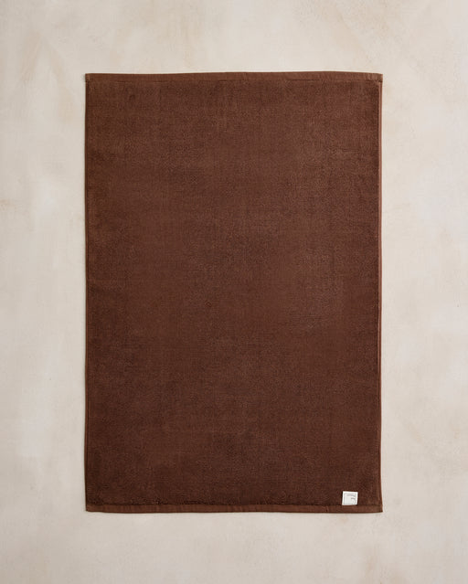 The Bed Threads Espresso Cotton Terry Bath Mat, large and rectangular, lies flat on a light beige surface with a small white fabric tag in the lower right corner.