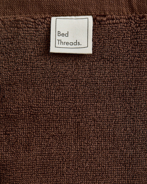 A close-up of the brown Espresso Cotton Terry Bath Mat by Bed Threads shows its textured, quick-drying fabric and a white Bed Threads. label sewn near the top.