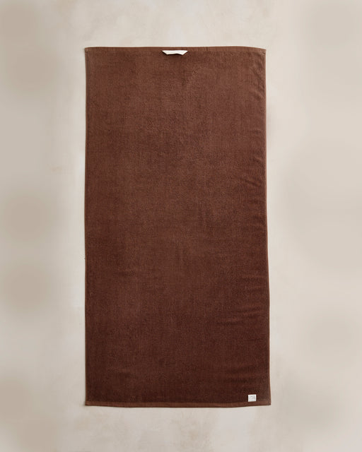 A set of two Bed Threads Espresso Cotton Terry Bath Towels hangs flat against a light beige wall, featuring a plush, dark brown finish with white tag and loop details. Highly absorbent, they’re ideal for everyday use.