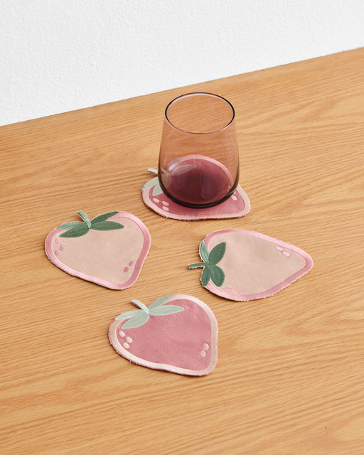 A glass of red wine rests on a Les Fruits Fraise Coaster by Bed Threads—strawberry-shaped pink clay—on a wooden table, with three more charming coasters from the set of four scattered nearby.