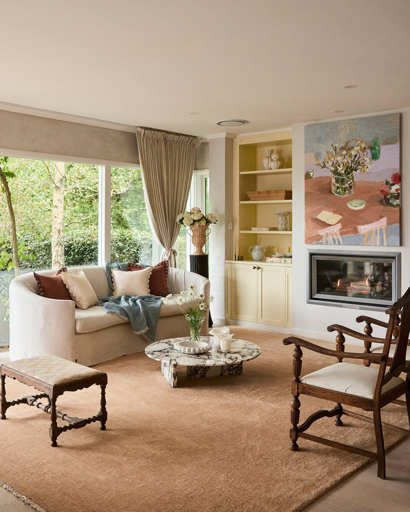 A cozy living room featuring a Bed Threads Lagoon & Crème 100% French Flax Linen Muslin Throw, a white linen sofa with pillows, marble coffee table with flowers, armchair, fireplace, art, decor shelves, and large windows overlooking green trees.