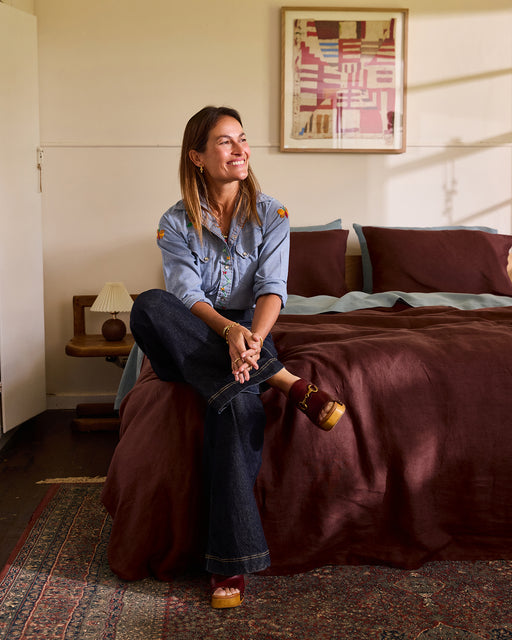 A smiling woman in a denim shirt and dark jeans sits on a maroon-covered bed made with the Bed Threads Cacao 100% French Flax Linen Bedding Set, in a cozy bedroom with a lamp on a wooden nightstand and abstract art above the bed.