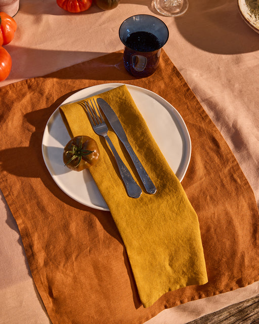 A white plate with a dark tomato sits atop a brown placemat, accompanied by a Bed Threads Turmeric 100% French Flax Linen Napkin, fork, and knife. A blue cup with a drink rests nearby as sunlight casts warm shadows over the scene.
