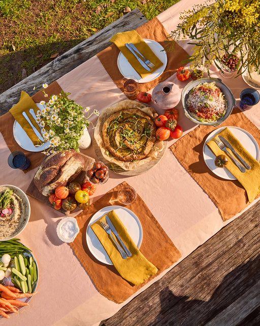 A picnic table outdoors is set with plates, bread, fresh vegetables, a tart, salad, flowers, and Bed Threads' Turmeric 100% French Flax Linen Napkins (Set of Four), all atop a striped pink and tan tablecloth in bright sunlight.