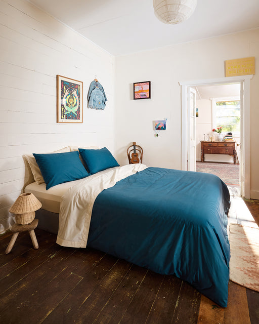 A cozy bedroom features a bed dressed in the Bed Threads Aegean Organic Cotton Duvet Cover, accented with blue and white pillows. Wooden floors, wall art, a lamp, and a guitar decorate the space. An open door reveals a sunlit room with a desk and plants.