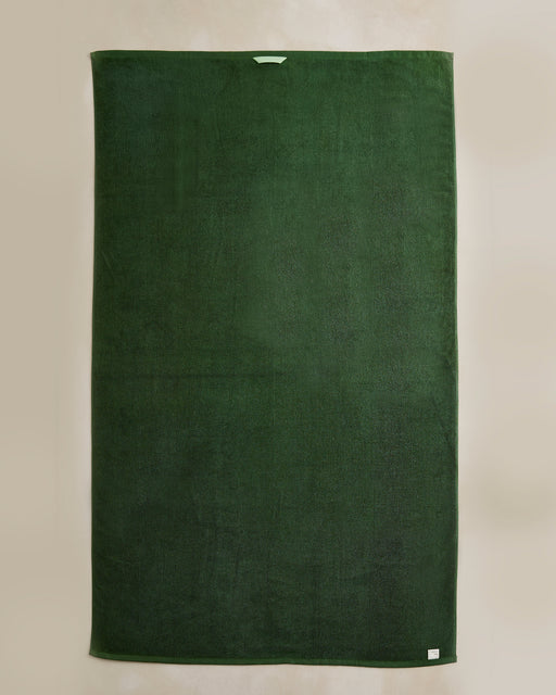 The Moss Cotton Terry Bath Sheet by Bed Threads, large and rectangular in dark green, is laid flat on a neutral background with a small white tag at one corner and a light loop at the top center.