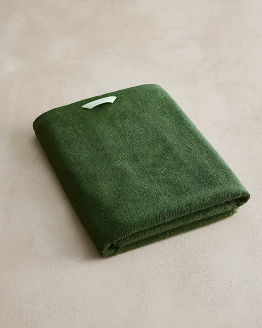 The Moss Cotton Terry Bath Sheet by Bed Threads, in dark green, is crafted from plush quick-dry cotton with a white tag and rests on a beige surface—ideal for luxe bathroom towel collections.