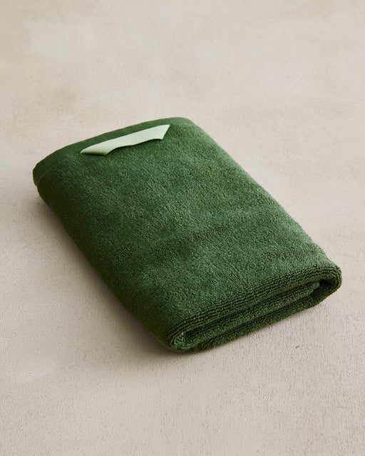 The Bed Threads Moss Cotton Terry Hand Towel, in dark green with a plush 650GSM texture and light green hanging loop, rests neatly folded on a beige surface.