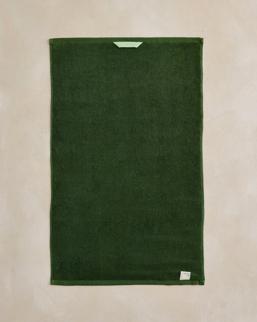 The Moss Cotton Terry Hand Towel by Bed Threads, in dark green, is laid flat on a beige surface. It features a small green hanging loop at the top and a white tag on one lower corner of its plush 650GSM cotton terry fabric.