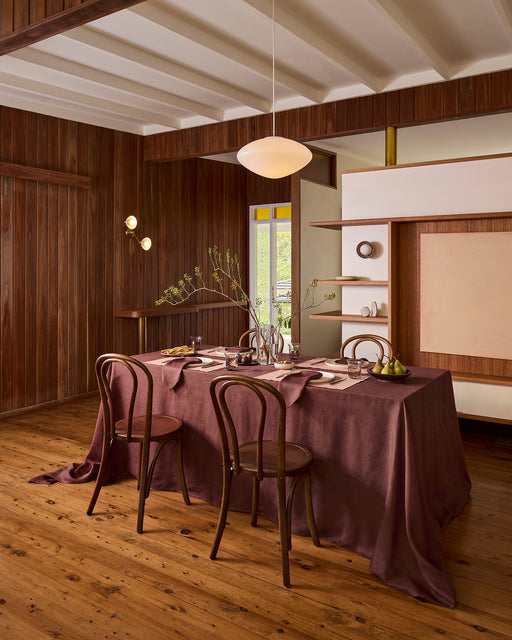 An outdoor meal is set with a brown tablecloth and a Lagoon 100% French Flax Linen Table Runner by Bed Threads, featuring bread, figs, grapes, cheese, placemats, white flowers in a vase, plates, silverware, and glasses.