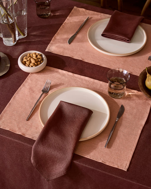A cozy dining room features wood-paneled walls and floors, a mauve-covered table with four wooden chairs, Bed Threads’ Hazelnut 100% French Flax Linen Placemats (Set of Four), simple tableware, pendant lighting, minimalist art, and tall branches in a vase.