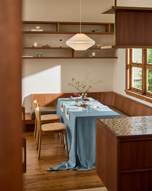 A dining table is set with a white plate, silverware, and a folded Lagoon 100% French Flax Linen Napkin by Bed Threads on a cream and blue tablecloth. Nearby is a plate of dessert, figs, and blackberries.