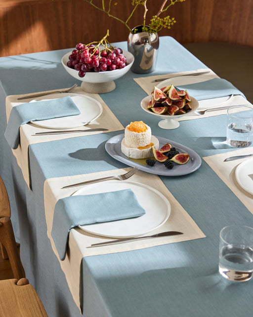 A dining table is set with Bed Threads’ Lagoon 100% French Flax Linen Napkins (Set of Four), plates, silverware, glasses of water, grapes in a bowl, sliced figs, berries, a coconut-topped dessert, and a silver vase with branches in the background.