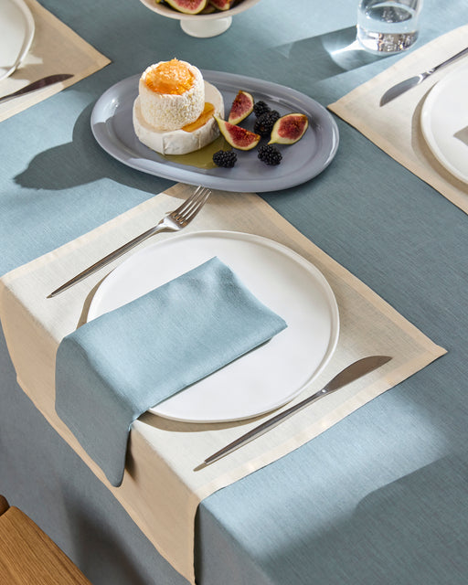 A modern dining room features natural wood furniture, built-in benches, open shelves, wooden floors, large windows, and a Bed Threads Lagoon 100% French Flax Linen Tablecloth draped over the table set for a meal.