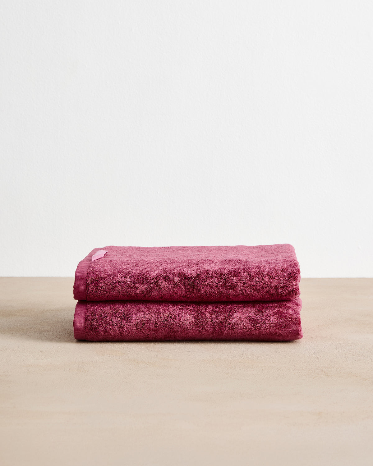 The Berry Cotton Terry Bath Towels (Set of Two) by Bed Threads are neatly folded, highly absorbent dark pink towels displayed on a light beige surface against a plain white background.