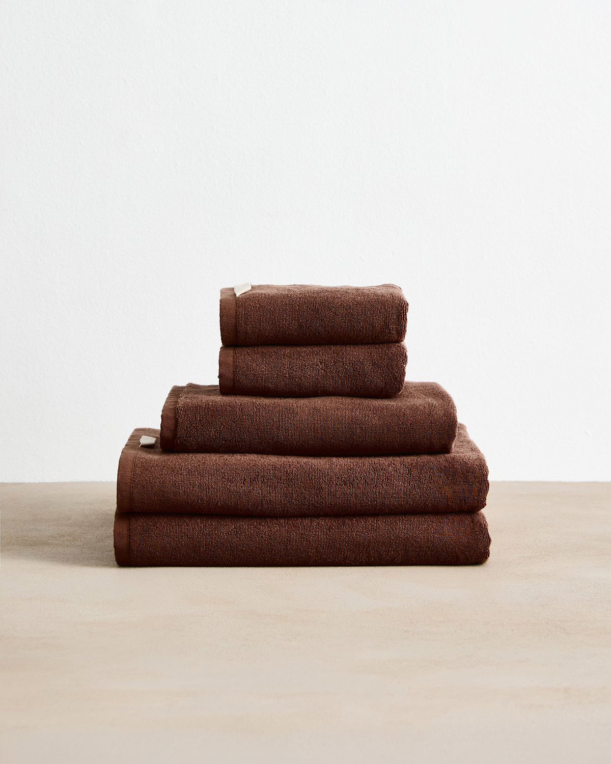 The Bed Threads Espresso Cotton Terry Bath Towel Bundle features four highly absorbent brown towels in assorted sizes, neatly stacked on a beige surface with a plain white background.