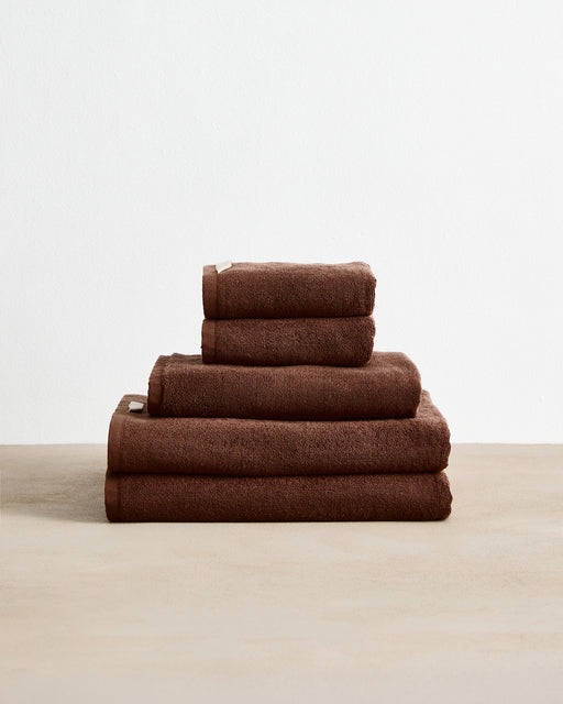 The Bed Threads Espresso Cotton Terry Bath Towel Bundle features four highly absorbent brown towels in assorted sizes, neatly stacked on a beige surface with a plain white background.