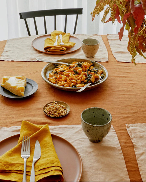 A table set with plates, yellow napkins, cutlery, ceramic cups, and pasta is elegantly protected by Bed Threads’ Terracotta 100% French Flax Linen Placemats (Set of Four), while cheese, pine nuts, and autumn leaves complete the scene.