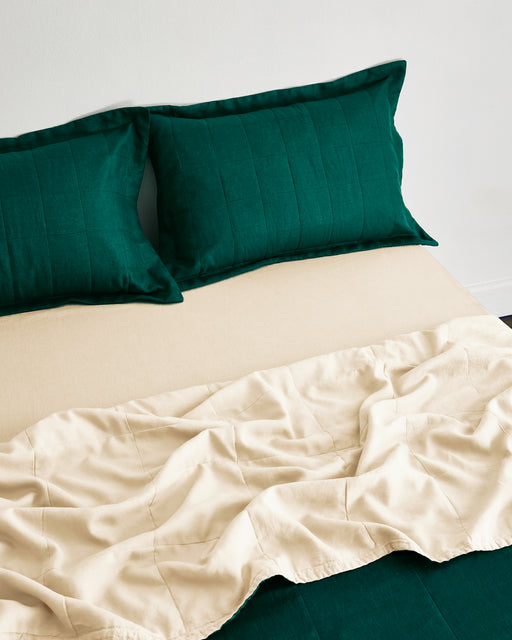 A neatly made bed with dark green pillows, a beige fitted sheet, and a partially rumpled beige blanket on top, set against a plain white wall.