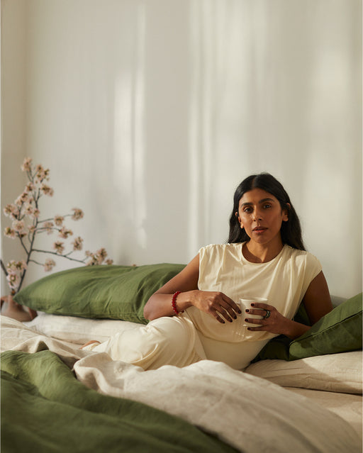 A woman in a light cream outfit reclines on a bed with green and beige bedding, resting against Bed Threads’ Olive 100% French Flax Linen Pillowcases. Sunlight filters in as delicate blossoms accent the peaceful scene.