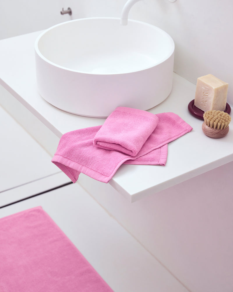 A modern white bathroom sink with a bar of soap, a brush, and two Bubblegum 100% Cotton Terry Face Washers by Bed Threads folded on the countertop. A matching pink bathmat completes the minimal, tidy scene designed to be gentle on skin.