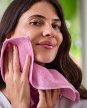 A woman with long brown hair smiles gently while holding a Bubblegum 100% Cotton Terry Face Washer (Bed Threads, set of two) to her cheek. The soft, absorbent fabric looks gentle on skin. The softly blurred background features green and neutral tones. - view