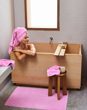 A person relaxes in a wooden tub with a pink Bubblegum 100% Cotton Terry Bath Sheet by Bed Threads wrapped around their head, plush quick-drying towels and a bath mat nearby, while a stool holds soap and water. - view