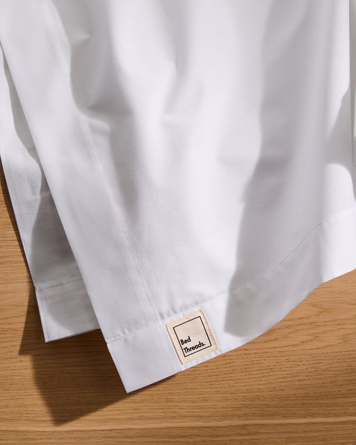 A close-up of the Crisp White Organic Cotton Flat Sheet by Bed Threads, folded on wood, shows a small Bed Threads fabric label near the hem—ideal breathable bedding that's gentle on skin.