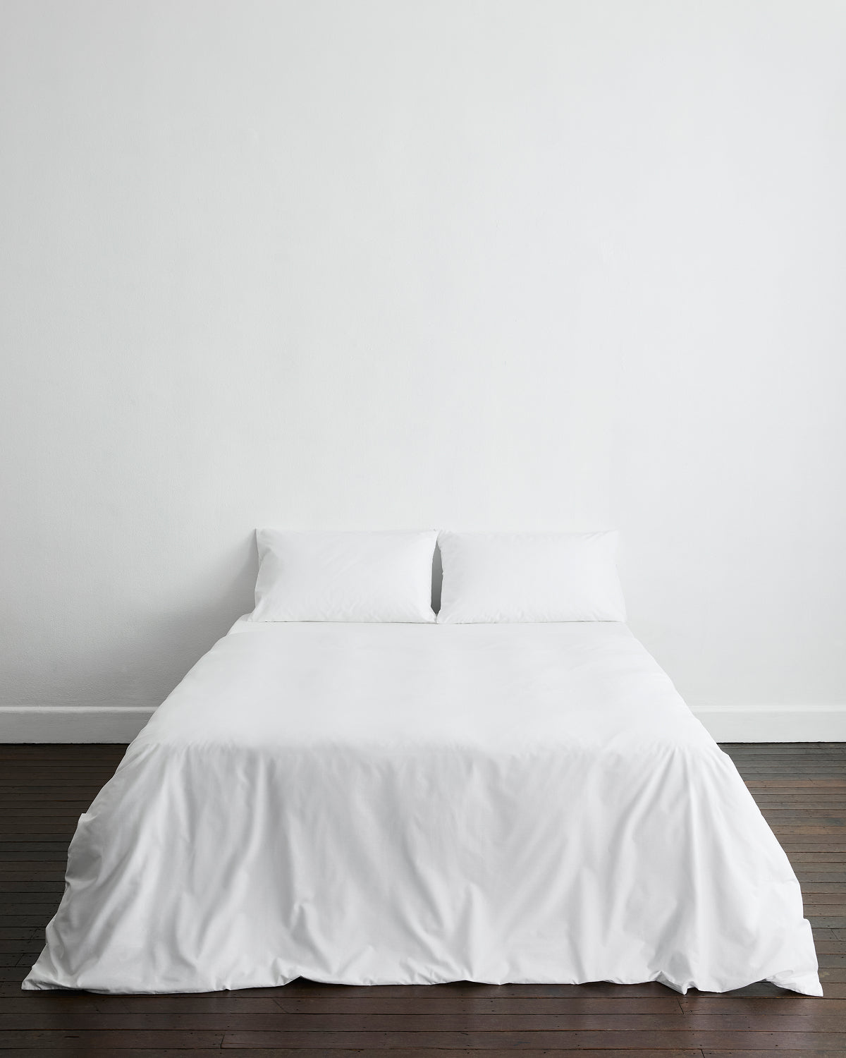 A minimalist room features a neatly made bed with white sheets and hotel-quality bedding, including the Bed Threads Crisp White Organic Cotton Duvet Cover, set against a plain white wall on a wooden floor.
