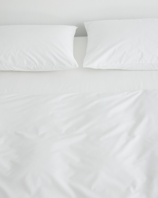 A neatly made bed viewed from above features Bed Threads' Crisp White Organic Cotton Pillowcases (Set of Two) and a white comforter, all set against a plain white wall in a minimalistic style.