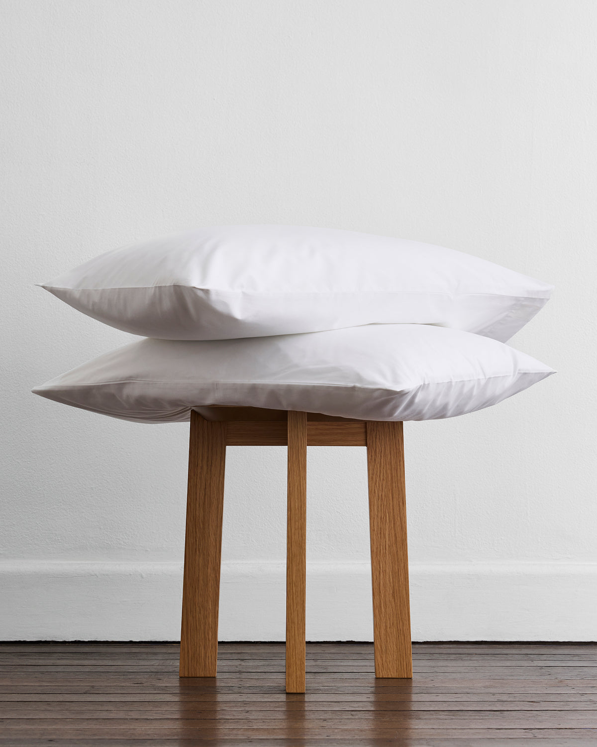 The Bed Threads Crisp White Organic Cotton Pillowcases are displayed on a simple wooden stool against a white wall, highlighting its fresh and natural aesthetic.