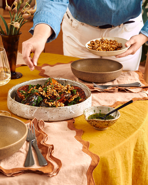 A person sprinkles chopped walnuts over roasted eggplant on a ceramic platter atop a yellow and peach cloth, with Bed Threads Terracotta & Rust French Flax Linen Scalloped Napkins, plates, cutlery, and a bowl of green sauce nearby.