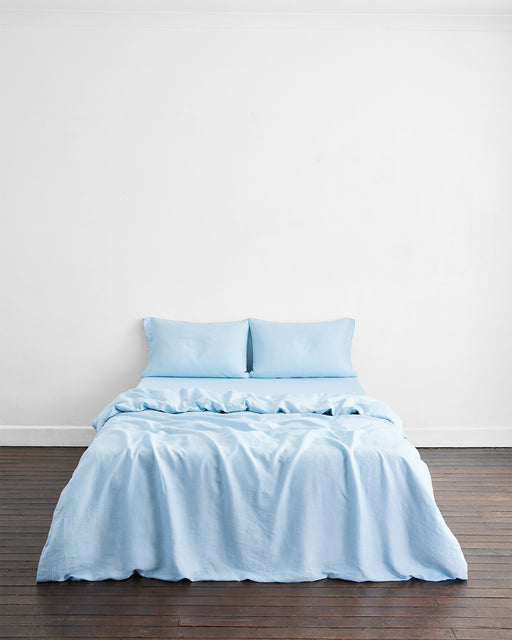 A neatly made bed with light blue sheets and two pillows sits against a white wall on a dark wooden floor, dressed in the Bed Threads Coast 100% French Flax Linen Duvet Cover, which looks soft and gently rumpled for a relaxed feel.