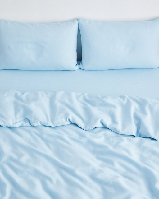 A bed features the Coast 100% French Flax Linen Duvet Cover by Bed Threads, slightly rumpled at the center, with light blue sheets and matching pillowcases; pillows are propped against a white wall.