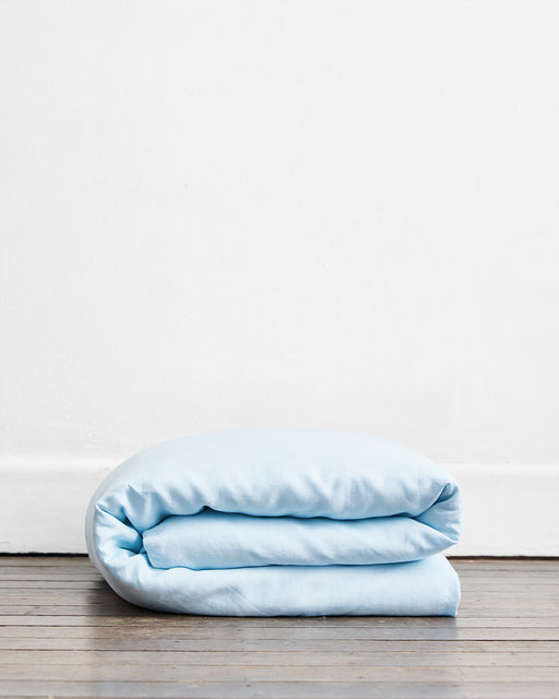 The Coast 100% French Flax Linen Duvet Cover by Bed Threads, in light blue, is neatly folded on a wooden floor against a plain white wall.
