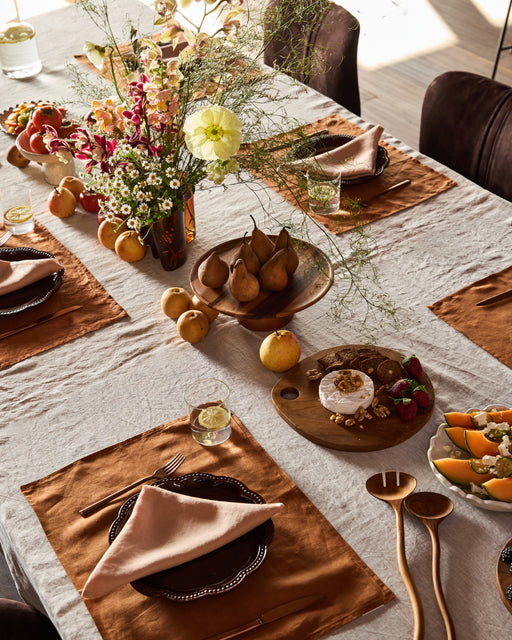 A dining table set for a meal features Bed Threads’ Terracotta 100% French Flax Linen Napkins (Set of Four), plates, wooden utensils, a floral centerpiece, fresh pears, a cheese board with nuts and fruit, and melon slices on a light tablecloth.