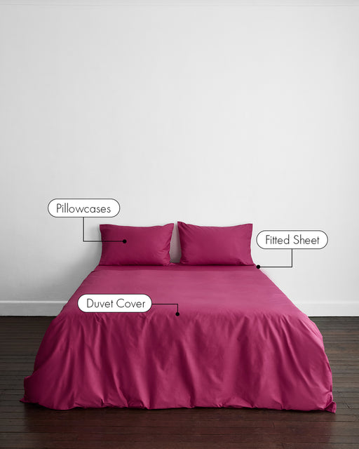 A bed dressed in the Bed Threads Berry Organic Cotton Bedding Set—two labeled pillowcases, fitted sheet, and duvet cover—appears against a plain white wall and a grayish floor.