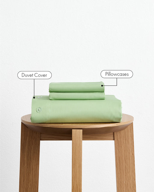 The Bed Threads Apple Organic Cotton Duvet Cover Set, including matching pillowcases, is neatly folded and stacked on a wooden stool with labels for each item, set against a plain white background.