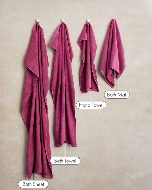 Four plush towels by Bed Threads—including a Berry Cotton Terry Bath Sheet—hang on hooks against a beige wall, each labeled for size and type, adding a touch of luxury to the bathroom.