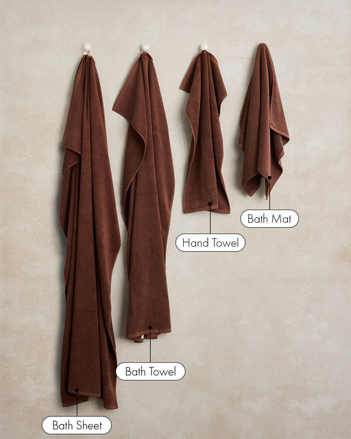 Two plush Espresso Cotton Terry Bath Towels by Bed Threads hang on wall hooks. Highly absorbent and clearly labeled, these towels stand out against a neutral beige wall.