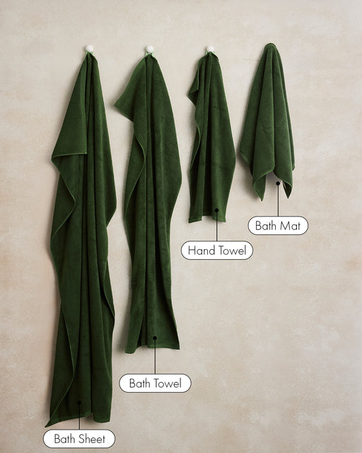 Four Moss Cotton Terry Hand Towels by Bed Threads, crafted from plush 650GSM cotton, hang on hooks against a beige wall. Each labeled: Bath Sheet (largest), Bath Towel, Hand Towel, and Bath Mat (smallest, square-shaped).