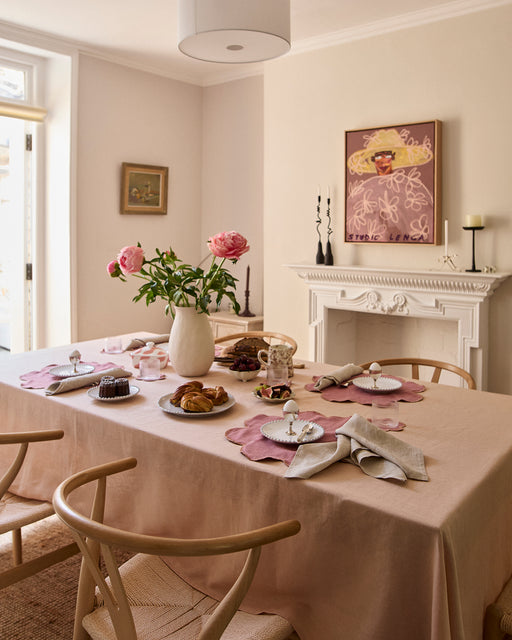 A dining room features wooden chairs, a light pink French flax linen tablecloth, and a table set for breakfast with pastries, fruit, and Bed Threads Pink Clay & Hazelnut 100% French Flax Linen Petal Placemats. Flowers in a vase add charm; natural light fills the space.