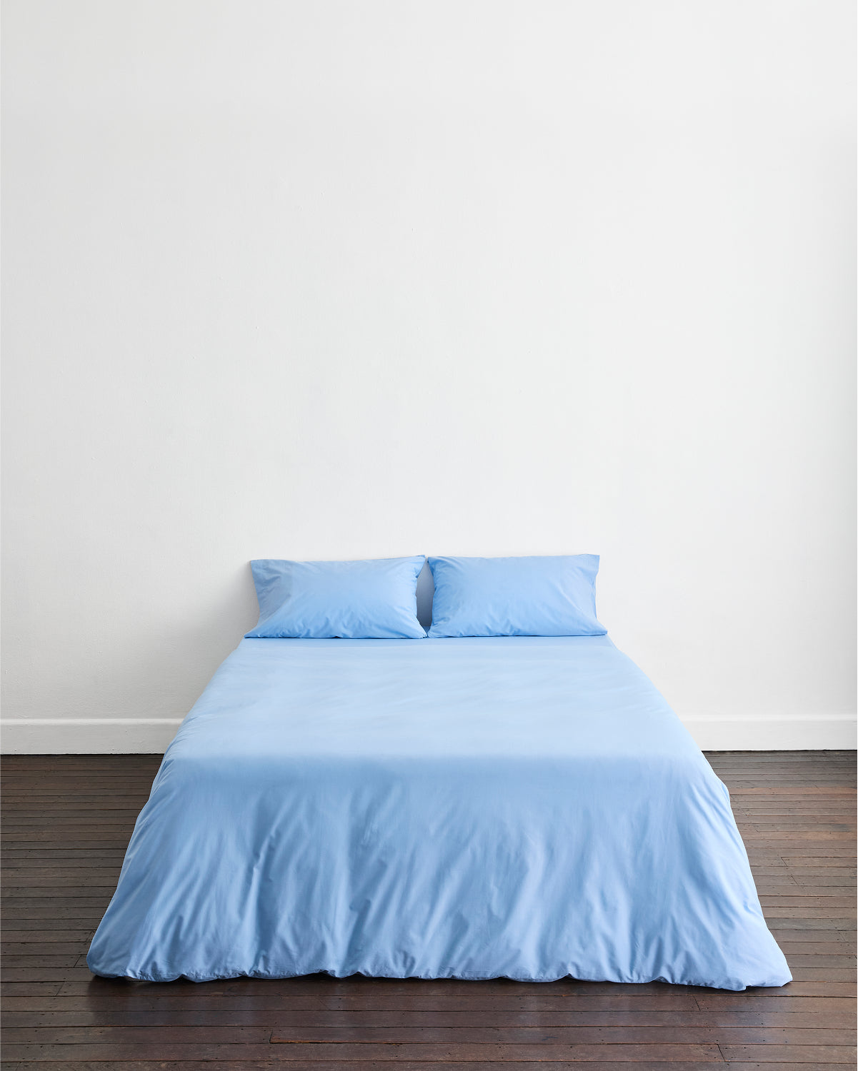 A neatly made bed features the Bed Threads Sky Organic Cotton Bedding Set and two pillows, set against a white wall on a dark wooden floor, highlighting the comfort of this percale bedding in a minimalistic room.