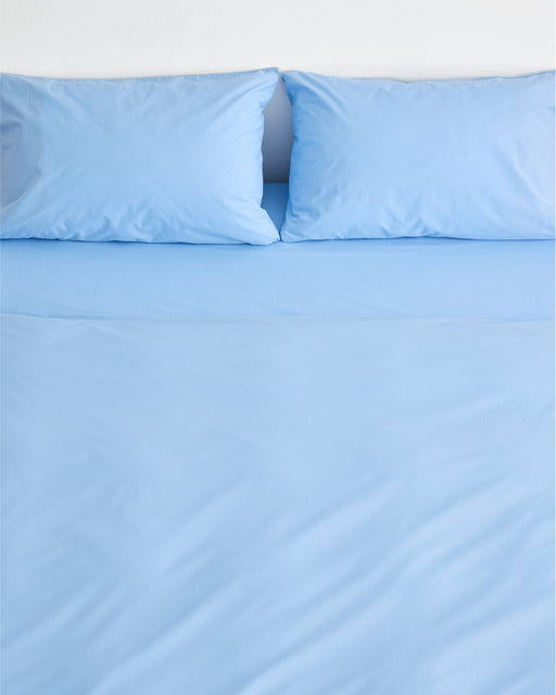 A neatly made bed features the Sky Organic Cotton Bedding Set by Bed Threads in light blue, with two matching pillows, set against a plain white wall.